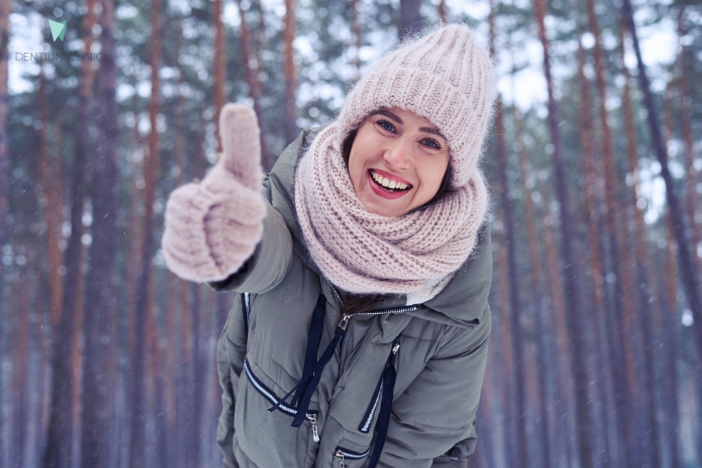 Low angle of cheerful smiling woman standing in the forest with thumb up. Giving thumb up t the camera while posing in the wintry forest